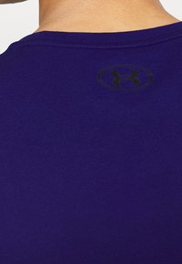 Närbild av en persons övre rygg som bär en mörkblå skjorta med en svart Under Armour-logotyp nära halsringningen.