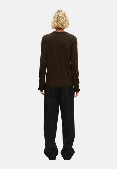 Persona con el cabello rizado y corto de color rubio, vistiendo una camiseta de manga larga marrón oscuro y pantalones negros holgados, de pie de espaldas sobre un fondo claro y liso.