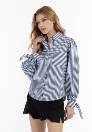 Pasiasta koszula button-up w niebiesko-białe paski, z kieszonką na piersi i zawiązywanymi mankietami, w połączeniu z czarnymi, postrzępionymi szortami. Lekki materiał bawełniany.