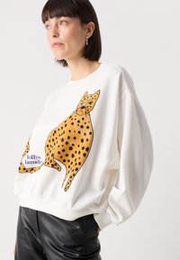 Felpa bianca con un'illustrazione grande di un leopardo arancione con macchie nere e occhi verdi, abbinata a pantaloni neri.