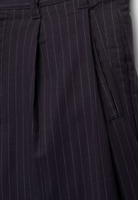 Pantalon bleu marine foncé à fines rayures blanches, avec des plis, des poches latérales et un tissu lisse et texturé. Coupe ajustée avec des détails subtils.