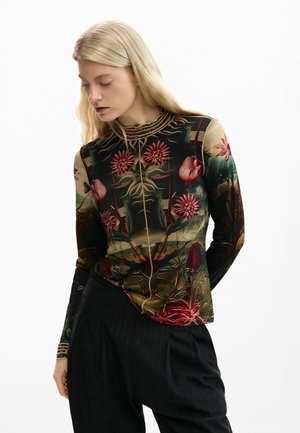 Mladá žena s dlhými blond vlasmi má na sebe priliehavé dlhé rukávy s detailným kvetinovým vzorom a voľné čierne nohavice, pozerá sa dolu.