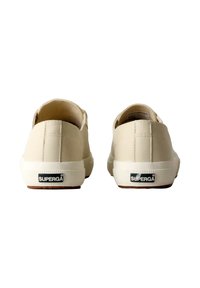 Zapatillas de lona beige con textura suave, de perfil bajo y suelas de goma blanca. Presentan un logo negro en el talón y detalles de costura.