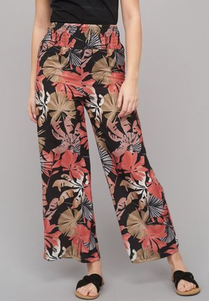 Woman wearing wide-leg pants with an elastic waistband, featuring a tropical floral print in red, beige, and black, paired with black slip-on sandals.