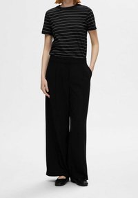 Schwarze weite Hosen mit hoher Taille, kombiniert mit einem schwarz-weißen gestreiften T-Shirt mit kurzen Ärmeln. Flache schwarze Schuhe runden das Outfit ab.