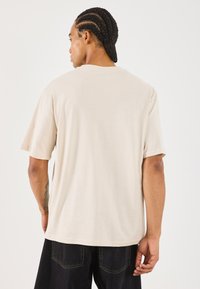 Beige T-Shirt mit kurzen Ärmeln, entspannter Passform, Rundhalsausschnitt und einfarbigem Textil, getragen über dunklen Jeans. Die Rückansicht zeigt schulterlanges Haar.