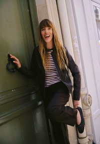 Femme aux cheveux longs portant un haut rayé et une veste noire souriante, tenant une poignée de porte noire d'une main et soulevant une chaussure noire de l'autre.