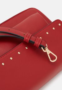 Angolo di borsa in pelle rossa con borchie dorate, tracolla rossa e nera e dettagli con clasp dorato su sfondo bianco.