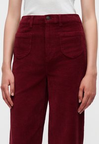 Gros plan d'une personne portant un pantalon en velours côtelé bordeaux taille haute avec des poches plaquées à l'avant et un top blanc sans manches côtelé.
