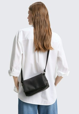 Schwarze, genarbte Leder-Crossbody-Tasche mit abnehmbarem Riemen, rechteckiger Form, mit einer Klappe verschlossen, die über einem weißen Hemd mit Knöpfen getragen wird.