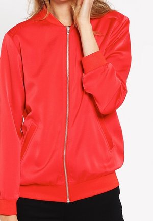 Femme portant une veste bomber rouge vif à fermeture éclair avec des poignets et une taille côtelés, touchant son visage d'une main.