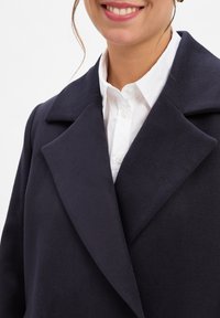 Marineblauw wollen blend jas met een gelapt kraag, met een zachte textuur en gestructureerde vorm. Witte blouse zichtbaar eronder.