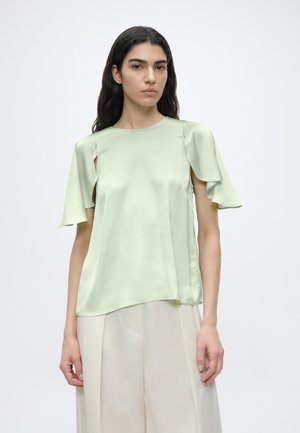 Femme aux longs cheveux foncés portant un chemisier en satin vert clair à manches volantées et un pantalon large couleur crème sur un fond uni.