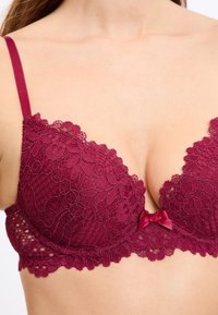 Bordowy koronkowy biustonosz z głębokim dekoltem, misternymi kwiatowymi wzorami, regulowanymi ramiączkami i małą satynową kokardką na środku.