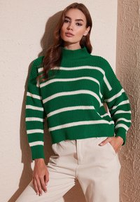 Maglione a righe verdi con collo alto, caratterizzato da strisce orizzontali bianche e una texture a coste; indossato con pantaloni chiari.