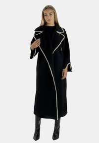 Lange zwarte jas met contrasterende witte stiksels, brede revers en uitlopende mouwen, gemaakt van een getextureerde stof, gedragen over kniehoge zwarte laarzen.