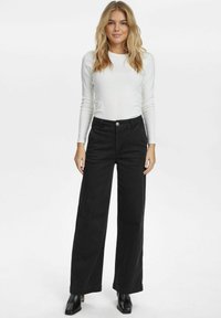Svarta vida jeans med hög midja och framknäppning, tillsammans med en figursydd vit långärmad skjorta och svarta ankelboots.
