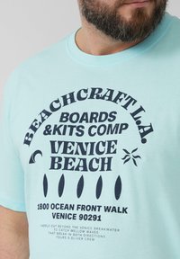 Lichtblauwe katoenen t-shirt met grote zwarte grafische tekst. Het ontwerp bevat "Beachcraft L.A." en "Venice Beach" met golf- en zonmotieven.
