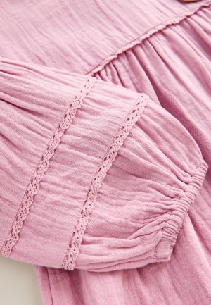 Secțiune de îmbrăcăminte roz cu mâneci strânse, având dantelă delicată la margine. Țesătura ușoară are un aspect moale, texturat.