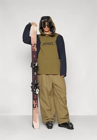 Isik, kellel on seljas oliivi- ja musta värvi suusajakk, laiad beežid püksid, mustad suusasaapad ja prillid, hoiab vertikaalselt käes roosa ja lilla suusapaari.