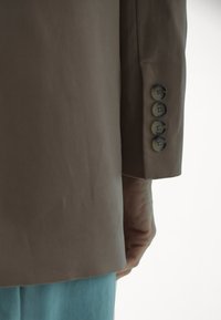 Barna kabátujj öt szövetből készült gombbal, egymás mellett; sima textúra és testhezálló fazon, világoskék nadrággal párosítva.