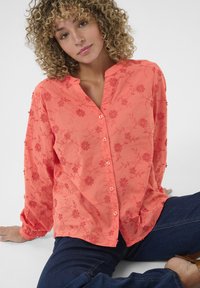 Blusa corallo con fodera a bottoni, caratterizzata da ricami floreali e una texture morbida, abbinata a jeans scuri e una vestibilità rilassata.