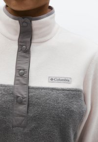 Artimos vaizdas dviejų tonų baltos ir pilkos spalvos vilnonio džemperio su pilkais spaudžiamais mygtukais ir Columbia logotipu ant krūtinės.
