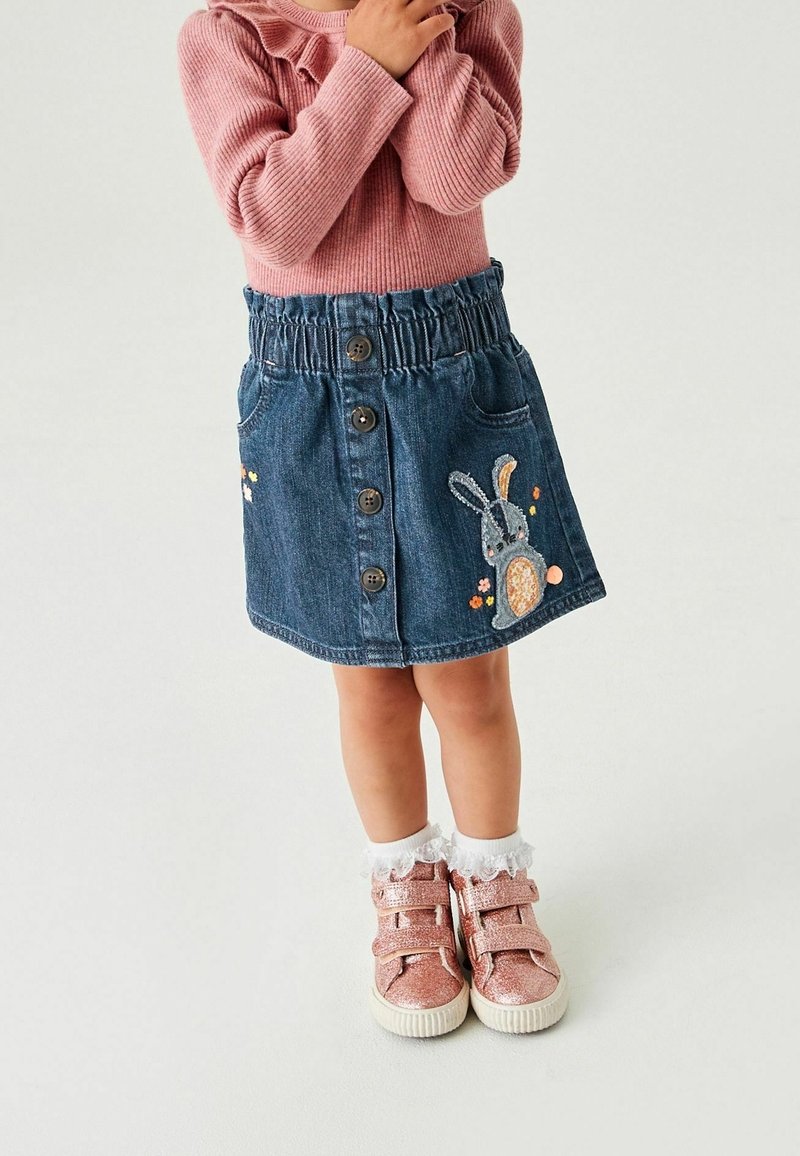 Jeune enfant portant un pull rose, une jupe en jean bleue avec des boutons à l'avant et un patch lapin, des chaussettes blanches en dentelle aux chevilles et des baskets roses pailletées.