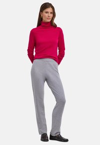 Dopasowany golf w jaskrawym różowym kolorze, zestawiony z jasnoszarymi, miękko teksturowanymi spodniami. Model stoi w czarnych mokasynach, ręce na biodrach.