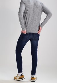 Grauer Strickpullover, dunkelblaue Jeans und braun-weiße Turnschuhe. Die Jeans haben einen Slim Fit und die Turnschuhe verfügen über ein auffälliges Logo auf der Rückseite.