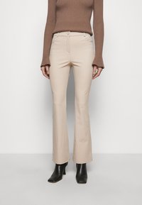 Pantalons évasés beiges avec une taille mi-haute, poches avant et design épuré, associés à des bottines noires, sur un fond uni.