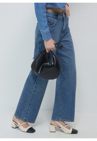 Sac hobo noir tenu à la main, porté avec un jean large en denim bleu et des escarpins à bride arrière crème avec des bouts noirs. Texture lisse, style décontracté.