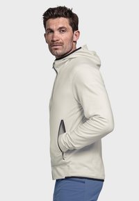 Hellbeige Zip-Jacke mit Reißverschluss und Kapuze, ausgestattet mit schwarzen Reißverschlussakzenten und Netz-Taschen. Hergestellt aus glattem, leichtem Material.