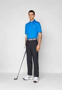 Blå polo skjorte med korte ermer, grå belte, svarte golfbukser og hvite golf sko. Står med en golfklubbe i hånden.
