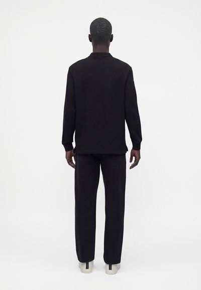 Personne aux cheveux courts portant un haut noir à manches longues, un pantalon noir et des baskets blanches, debout de dos sur un fond blanc.