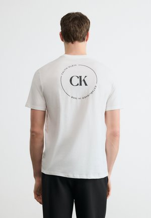 Άνδρας που φοράει λευκό κοντομάνικο T-shirt με μαύρο λογότυπο "CK" και κυκλικό κείμενο "Calvin Klein Established in 1968" στην πίσω πλευρά, μαύρο παντελόνι.