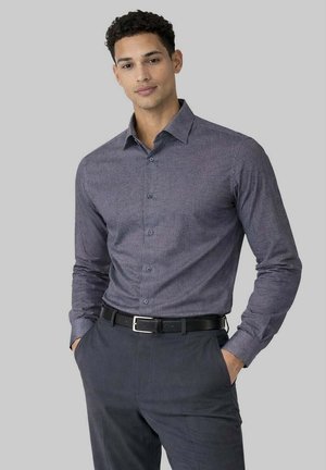 Homme portant une chemise grise à manches longues boutonnée, rentrée dans un pantalon gris foncé avec une ceinture noire, les mains dans les poches, debout devant un fond clair uni.