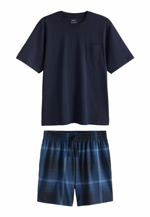 Man staat blootsvoets in een kamer, draagt een marineblauw T-shirt en blauwe geruite korte broek, met één hand in de zak en grijze muren op de achtergrond.