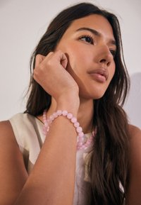 Rosa Perlenarmband aus glatten Steinen mit runden Perlen. Die Trägerin trägt eine passende rosa Perlenkette und ein neutrales Oberteil.