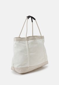 Tote-väska i ljuskräm canvas med en vadderad botten. Funktioner två rep-handtag, ett med en svart accent, som visar en elegant, minimalistisk design.