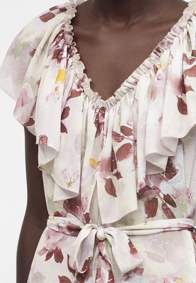 Άτομο με σκουρόχρωμο δέρμα που φοράει λευκό ανθισμένο φόρεμα με σούρες στη λαιμόκοψη και κοντά φουσκωτά μανίκια, δεμένο στη μέση με ζώνη από το ίδιο ύφασμα.