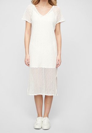 Femme portant une robe midi en maille blanche à manches courtes avec un col en V et des baskets blanches, debout devant un fond gris uni.