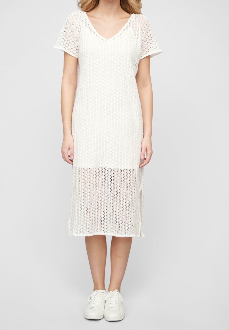 Femme portant une robe midi en maille blanche à manches courtes avec un col en V et des baskets blanches, debout devant un fond gris uni.