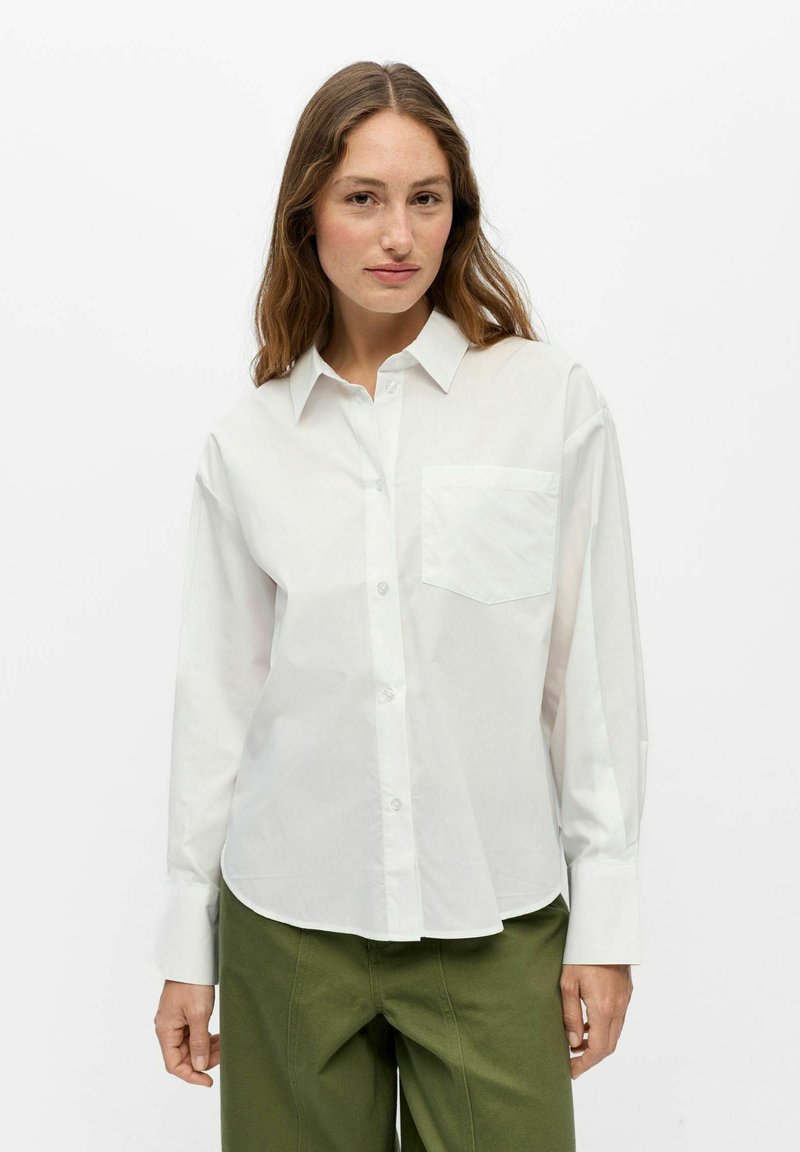 Femme aux longs cheveux bruns portant une chemise blanche boutonnée et un pantalon vert olive, debout devant un fond blanc uni.