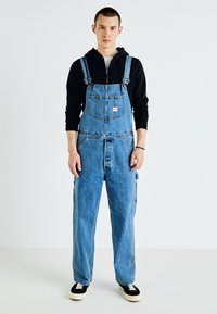 Light blue denim overalls with adjustable straps and front pockets, worn over a black hoodie. Model stands against a plain background.