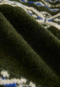 Strikket uldstof i mørkegrøn med et blåt og hvidt geometrisk mønster, der fremviser en blød, tekstureret overflade med synlige fibre.