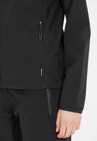 Veste softshell noire avec zippers, au design épuré et branding minimal. Présente une coupe ajustée et une texture lisse.