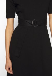 Schwarzes, strukturiertes Kleid mit tailliertem Schnitt, kurzen Ärmeln und einem breiten Gürtel mit einer gravierten Schnalle, das ein minimalistisches Design präsentiert.