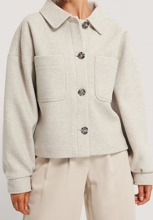 Lyse beige uldjakke med knapper, store forlommer, bred krave og lange ærmer, båret med matchende beige bukser.