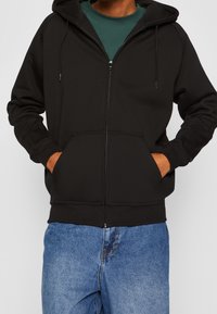 Sudadera negra con cremallera y capucha ajustable, bolsillos frontales y puños de canalé. Llevada sobre una camiseta verde y combinada con vaqueros azules.
